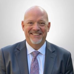a Smiling Man in a Suit and Tie Bald with a Goatee Against a Light Background