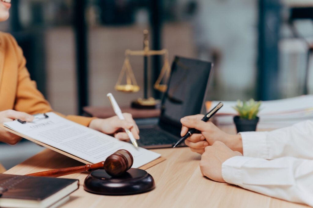Two Professionals Signing a Contract at a Desk with a Gavel and Scales of Justice Indicating a Legal Agreement